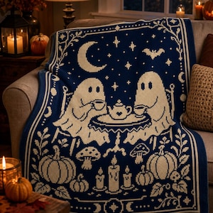 May include: A dark blue knit throw blanket featuring a cream-colored pattern of two ghosts sitting at a table drinking tea. The design includes a crescent moon, stars, a bat, pumpkins, mushrooms, and candles, all framed by a decorative border.