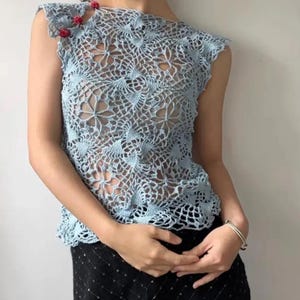 May include: A light blue crochet top with a floral pattern. The sleeveless top features a delicate openwork design and is adorned with small red embellishments on the shoulder. The top is paired with a black skirt.