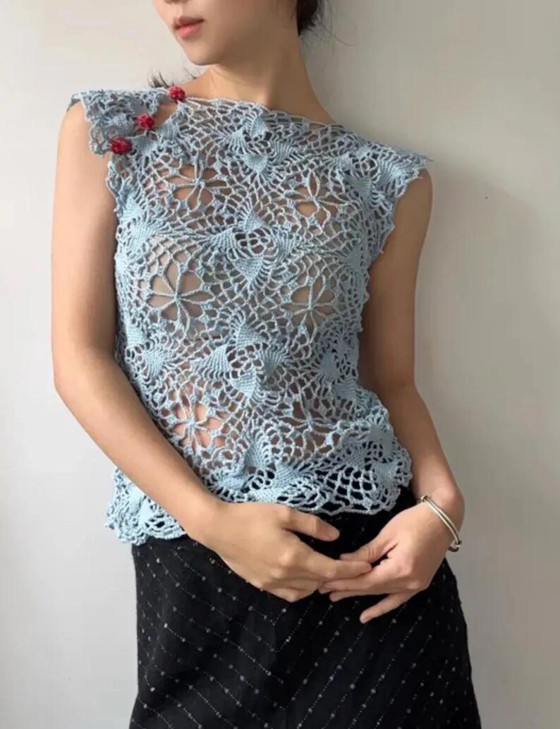 May include: A light blue crochet top with a floral pattern. The sleeveless top features a delicate openwork design and is adorned with small red embellishments on the shoulder. The top is paired with a black skirt.