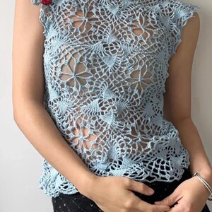 May include: A light blue crochet top with a floral design. The sleeveless top features a delicate openwork pattern and is adorned with small red embellishments on the shoulder. The top is paired with a black skirt.