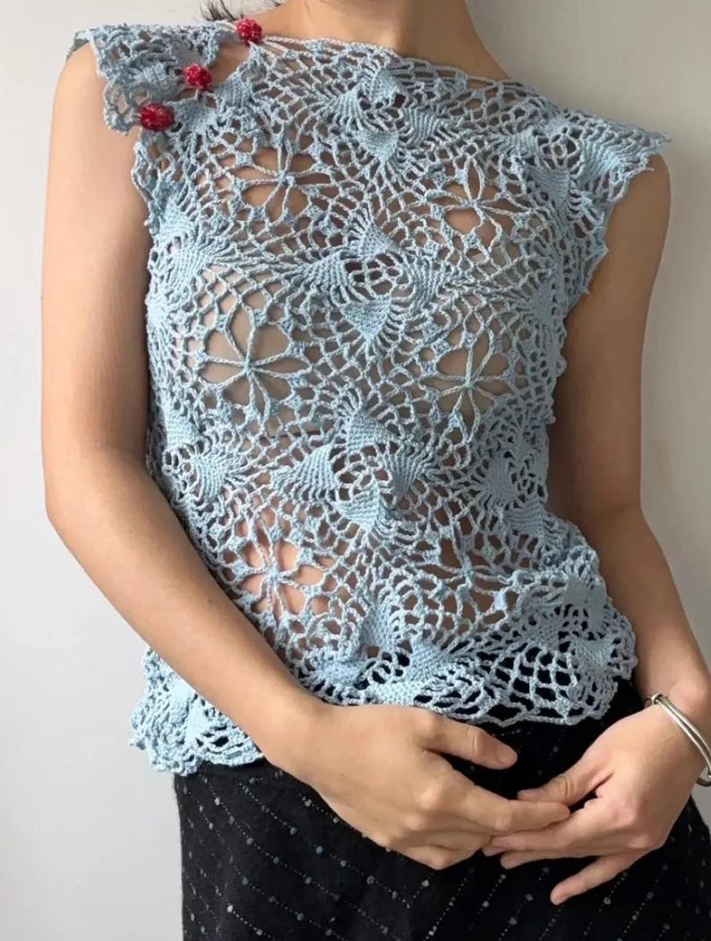 May include: A light blue crochet top with a floral design. The sleeveless top features a delicate openwork pattern and is adorned with small red embellishments on the shoulder. The top is paired with a black skirt.