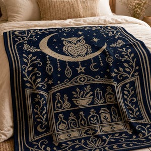 May include: A dark blue woven throw blanket featuring a cream-colored pattern of an owl perched on a crescent moon, surrounded by stars, moths, and hanging crystals. Below, a cabinet displays various potion bottles, plants, and crystals.