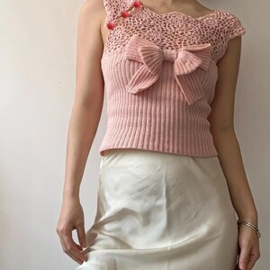 May include: Pink crochet top with a bow detail and ribbed knit bodice. The top features a delicate crochet neckline and is adorned with small red embellishments. A light pink top, perfect for a summer outfit.