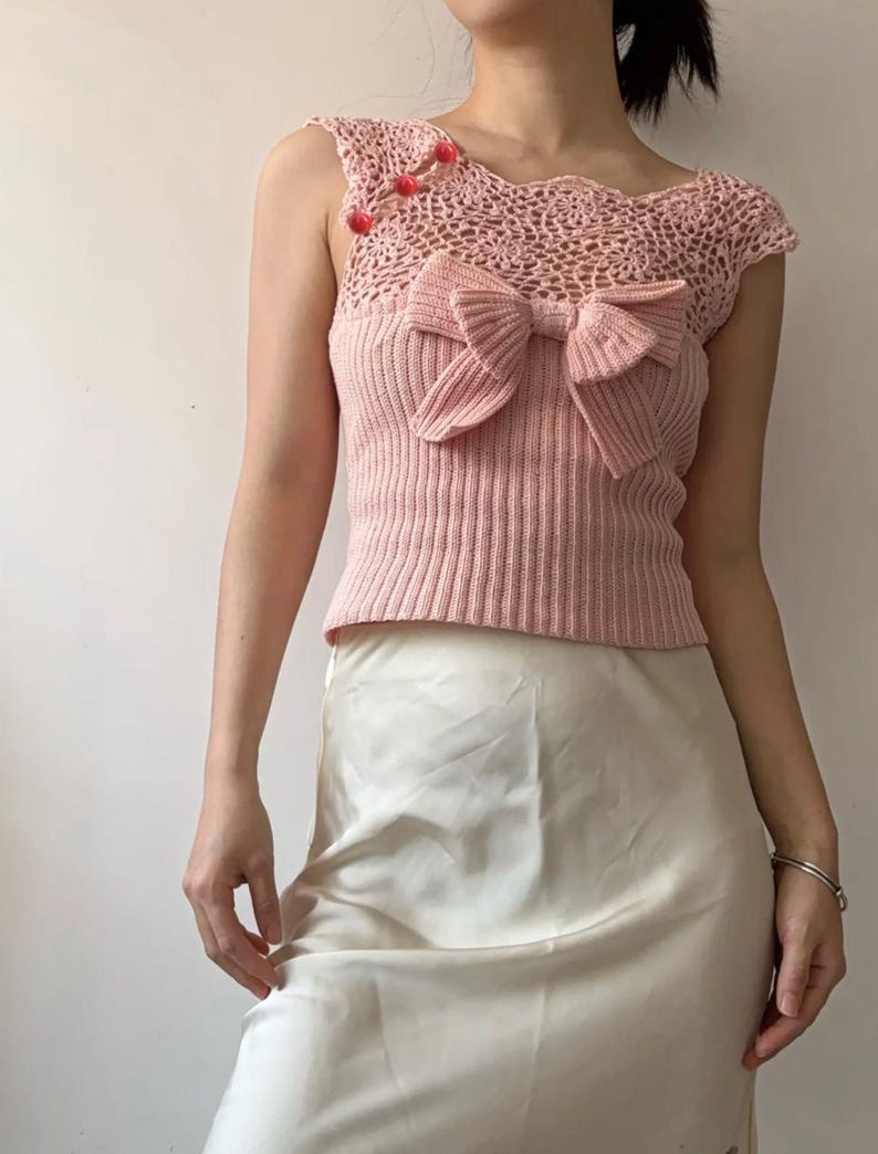 May include: Pink crochet top with a bow detail and ribbed knit bodice. The top features a delicate crochet neckline and is adorned with small red embellishments. A light pink top, perfect for a summer outfit.