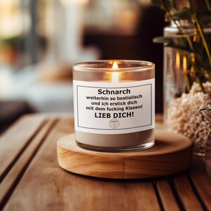 May include: A lit candle in a clear glass jar with a gray base, sitting on a wooden coaster. The candle has a white label with black text that reads "Schnarch weiterhin so bestialisch und ich erstick dich mit dem fucking Kissen! LIEB DICH!"