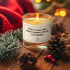 Könnte beinhalten: Eine brennende Kerze in einem durchsichtigen Glas mit einem weißen Etikett mit der Aufschrift "This is your gift. Don't expect more, I'm broke!" Die Kerze steht auf einer Holzoberfläche mit Tannenzapfen und Weihnachtsdekorationen.