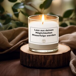 May include: A lit candle in a clear glass jar with a white label and gray base, sitting on a wooden slice. The label has German text. The background is blurred with green foliage and a brown cloth.