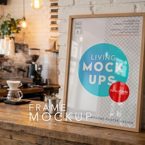 May include: A framed poster mockup with the text "LIVING MOCKUPS" in a cafe setting. The frame is light brown, and the poster has a blue circle and a red circle. The cafe has a coffee machine and other coffee-making equipment.