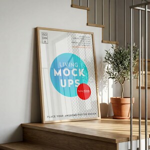 Op de afbeelding: Een ingelijste poster-mockup die tegen een muur op houten trappen leunt. De poster toont de tekst "LIVING MOCK UPS" in een blauwe cirkel, met daaronder een rode cirkel. Een kleine potplant is ook zichtbaar.
