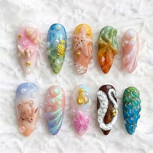 May include: A collection of ten artificial nails with a sea life theme. Each nail features a unique design, including seashells, starfish, and sea turtles, with colors like pink, blue, and gold. The nails are oval-shaped.