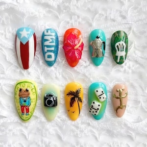 May include: A collection of ten artificial nails, each uniquely designed. The designs include a Puerto Rican flag, the letters "OTMF", a red hibiscus flower, a silver coffee pot, a green chair, a frog, a camera, a palm tree, dominoes, and a gold symbol.