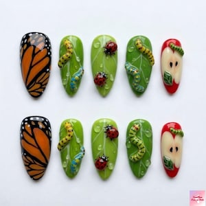May include: A set of ten artificial nails with nature-themed designs. The designs include a monarch butterfly wing, green leaves with ladybugs and caterpillars, and apple slices with worms. The nails are painted in various colors.