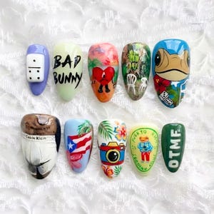 Op de afbeelding: Een verzameling van tien beschilderde kunstnagels. Ontwerpen zijn onder meer een domino, de woorden "BAD BUNNY", een hartvormige figuur, een tropische scène, een kikker, een Calvin Klein-ontwerp, een Puerto Ricaanse vlag, een camera, een kikker met tekst en de letters "OTMF".