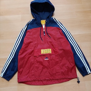 Op de afbeelding: Rode en marineblauwe Adidas anorakjas met capuchon. De jas heeft witte strepen op de mouwen en een voorzak met de tekst "WORLD CUP" en het Adidas-logo. De jas heeft een halve ritssluiting.