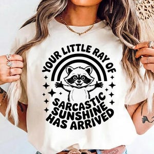 May include: Cream-colored t-shirt featuring a cartoon raccoon with outstretched arms, set against a rainbow and stars. The text reads "YOUR LITTLE RAY OF SARCASTIC SUNSHINE HAS ARRIVED" in a playful, bold font. A fun, casual graphic tee.