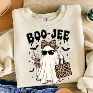 Könnte beinhalten: Ein cremefarbenes Sweatshirt mit Rundhalsausschnitt und einem Grafikdruck eines Geistes mit schwarzer Sonnenbrille und einer Schleife mit Leopardenmuster. Der Geist hält einen passenden Kaffeebecher und eine Tasche, mit dem Text BOO-JEE in schwarzer Tropfschrift.