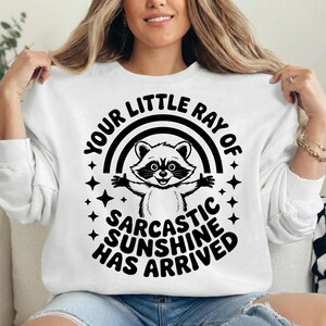 May include: White sweatshirt with a black graphic of a raccoon under a rainbow and the text "YOUR LITTLE RAY OF SARCASTIC SUNSHINE HAS ARRIVED". The raccoon has its arms up and is surrounded by stars.