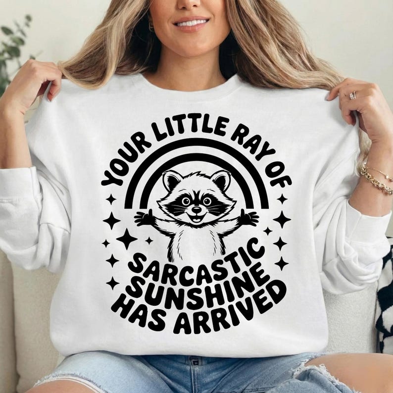 May include: White sweatshirt with a black graphic of a raccoon under a rainbow and the text "YOUR LITTLE RAY OF SARCASTIC SUNSHINE HAS ARRIVED". The raccoon has its arms up and is surrounded by stars.