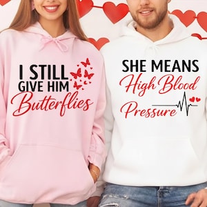 May include: Two matching hoodies, one pink and one white. The pink hoodie says "I STILL GIVE HIM Butterflies" with butterfly graphics. The white hoodie says "SHE MEANS High Blood Pressure" with a heartbeat graphic.