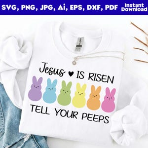 May include: White crew neck sweatshirt with the text "Jesus is Risen, Tell Your Peeps" and colourful Easter bunny graphics. A silver necklace rests on the shirt. The top is folded on a denim surface.