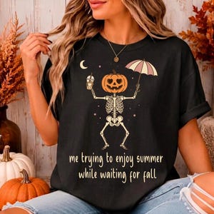 Pode incluir: Uma t-shirt preta de manga curta com um gráfico de um esqueleto com cabeça de abóbora segurando uma bebida e um guarda-chuva. O design inclui uma lua crescente e estrelas. O texto diz: me trying to enjoy summer while waiting for fall.