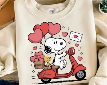 Könnte beinhalten: Cremefarbenes Sweatshirt mit einer Cartoon-Figur von Snoopy, die einen roten Roller fährt und herzförmige Ballons hält. Ein Korb mit Herzen und Woodstock ist ebenfalls auf dem Roller zu sehen. Ein weißer Umschlag mit einem Herz ist ebenfalls sichtbar.