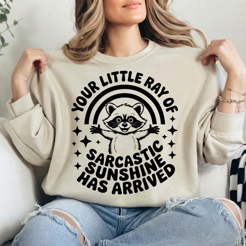 May include: A beige sweatshirt with the text "YOUR LITTLE RAY OF SARCASTIC SUNSHINE HAS ARRIVED" in black. The graphic features a raccoon with a rainbow and stars.