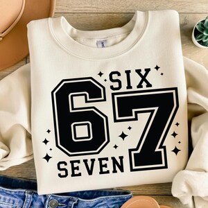 May include: Cream-colored sweatshirt with the number "67" in a black, collegiate-style font. The word "SIX" is above the number, and "SEVEN" is below. Small black star graphics are scattered around the numbers and words.