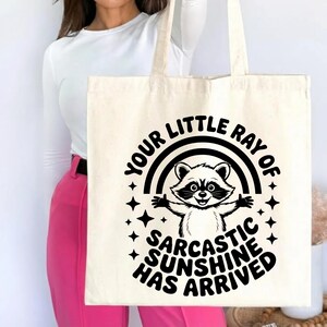 May include: Cream-colored tote bag featuring a black and white raccoon illustration with a rainbow and stars. The text reads "YOUR LITTLE RAY OF SARCASTIC SUNSHINE HAS ARRIVED" in a playful font. The bag has two handles.