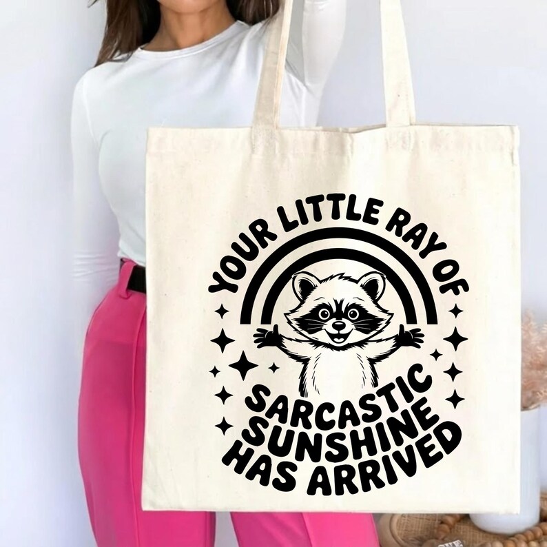 May include: Cream-colored tote bag featuring a black and white raccoon illustration with a rainbow and stars. The text reads "YOUR LITTLE RAY OF SARCASTIC SUNSHINE HAS ARRIVED" in a playful font. The bag has two handles.