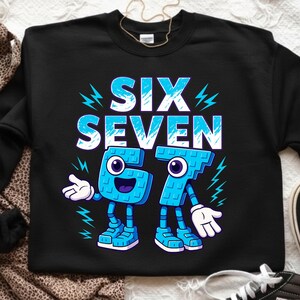 May include: Black sweatshirt with the words "SIX SEVEN" in large, light blue letters. Below the words are the numbers 6 and 7, designed as cartoon characters with arms, legs, and smiling faces. Lightning bolt graphics are on the sides.