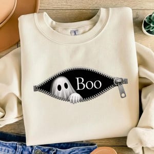 May include: A cream-colored crewneck sweatshirt featuring a graphic of a black oval shape with a silver zipper border. Inside the opening, a white ghost with two black eyes and a hand is visible next to the white text that reads Boo.