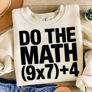 Può includere: Felpa color crema con la scritta "DO THE MATH (9x7)+4" stampata in grassetto nero. La felpa ha un girocollo e maniche lunghe. L'immagine è scattata su una superficie di legno con un paio di jeans blu e un cappello sullo sfondo.