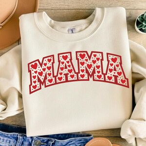 Könnte beinhalten: Cremefarbenes Sweatshirt mit dem Wort "MAMA" in roten, herzgefüllten Buchstaben. Die Buchstaben sind rot umrandet und mit kleinen roten Herzen und weißen Punkten gefüllt. Das Sweatshirt ist aus weichem Material.