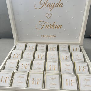 Könnte beinhalten: Eine offene, weiße Schachtel gefüllt mit kleinen, quadratischen Pralinen. Der Deckel trägt die Namen "Ilayda" und "Furkan" in goldener Schrift, ein Herz und das Datum 14.02.2026. Jede Pralinenverpackung zeigt die Initialen "IF".