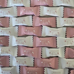 Könnte beinhalten: Nahaufnahme von Reihen verpackter Süßigkeiten in Rosa und Weiß. Jeder Wickler ist mit dem Namen "Elif", dem Wort "HOŞGELDIN" und Herzmotiven bedruckt. Die Süßigkeiten sind in einem Raster angeordnet und erzeugen einen ansprechenden visuellen Effekt.