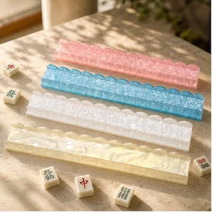 May include: Four rectangular mahjong tile racks with scalloped top edges, featuring a marbled finish in pink, blue, white, and cream. Each rack is positioned horizontally, accompanied by several scattered white mahjong tiles with red and green symbols.