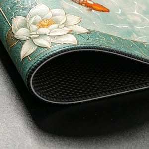May include: A decorative mousepad featuring a serene aquatic scene. The design includes a white lotus flower, a koi fish, and water elements in shades of teal and gold. The mousepad has a black, textured underside and a stitched edge.