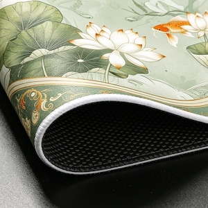 May include: A rectangular desk mat featuring a light green background with white lotus flowers, large green lily pads, and an orange koi fish. The mat has a white stitched edge and a black textured rubber base for grip on the underside.