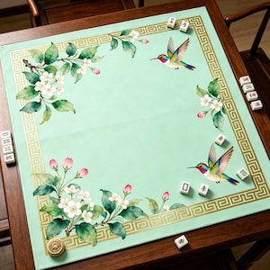 Puede incluir: Un tapete de juego de Mahjong verde claro con un borde dorado e ilustraciones florales y de colibríes. El tapete está sobre una mesa de madera oscura, con fichas de Mahjong esparcidas por la superficie.