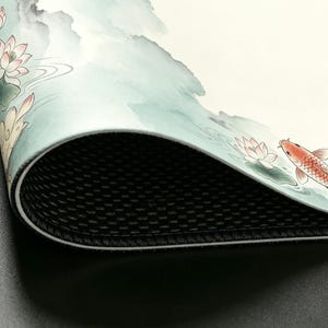 May include: A close-up of a decorative mat with a water color design. The mat features a koi fish, lotus flowers, and water elements in shades of red, pink, and green. The mat has a black, textured underside.