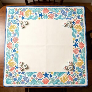 Pode incluir: Um tapete de mahjong quadrado com um centro creme e uma borda com ilustrações coloridas de conchas, ondas azuis, coral amarelo, estrelas-do-mar e cavalos-marinhos. Pequenas peças brancas de mahjong estão colocadas perto dos quatro cantos.