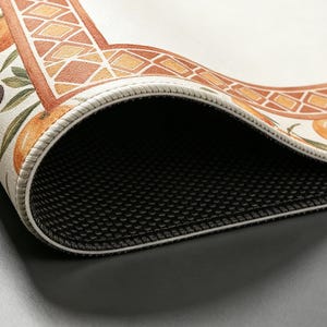 May include: Close-up of a decorative rug with a detailed border featuring geometric patterns in orange and brown. The rug's design includes illustrations of oranges and olive branches. The underside of the rug is black and textured.