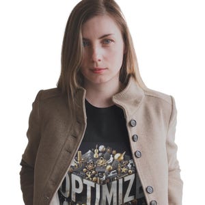 Pode incluir: Uma mulher usando um casaco bege sobre uma camiseta preta. A camiseta apresenta um gráfico 3D com a palavra "OPTIMIZE" e vários objetos em miniatura. O casaco tem um design abotoado.