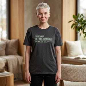 Puede incluir: Una camiseta gris oscuro con el texto "HG RUN_COMMAND" en blanco. Texto adicional está presente arriba y abajo del texto principal. La persona lleva pantalones negros.
