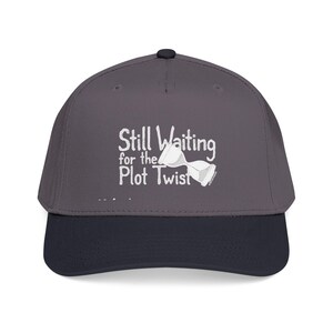 Puede incluir: Gorra de béisbol gris con visera azul marino. La gorra presenta el texto blanco "Still Waiting for the Plot Twist" y un gráfico de reloj de arena. Tiene una visera curva y un botón en la parte superior.