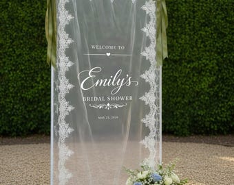 May include: A rectangular white metal frame holding a sheer fabric panel with white lace trim. The fabric features the text "WELCOME TO Emily's BRIDAL SHOWER MAY 25, 2026". Two olive green ribbons are tied to the top corners, and a floral arrangement sits at the base.