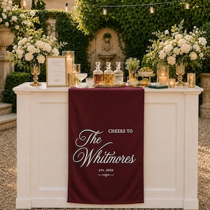 May include: A deep burgundy fabric table runner featuring white printed text that reads Cheers to The Whitmores Est. 2026, draped over a white rectangular bar counter. The runner is centered on the front panel of the bar, which is decorated with floral arrangements, glass decanters, and candles.