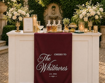 May include: A deep burgundy fabric table runner featuring white printed text that reads Cheers to The Whitmores Est. 2026, draped over a white rectangular bar counter. The runner is centered on the front panel of the bar, which is decorated with floral arrangements, glass decanters, and candles.