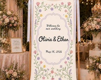 May include: A vertical cream-coloured fabric wedding welcome sign featuring a decorative purple border and floral illustrations. The text reads Welcome to our wedding, Olivia & Ethan, and May 18, 2026. The banner is suspended from a wooden bar with light grey ribbons.